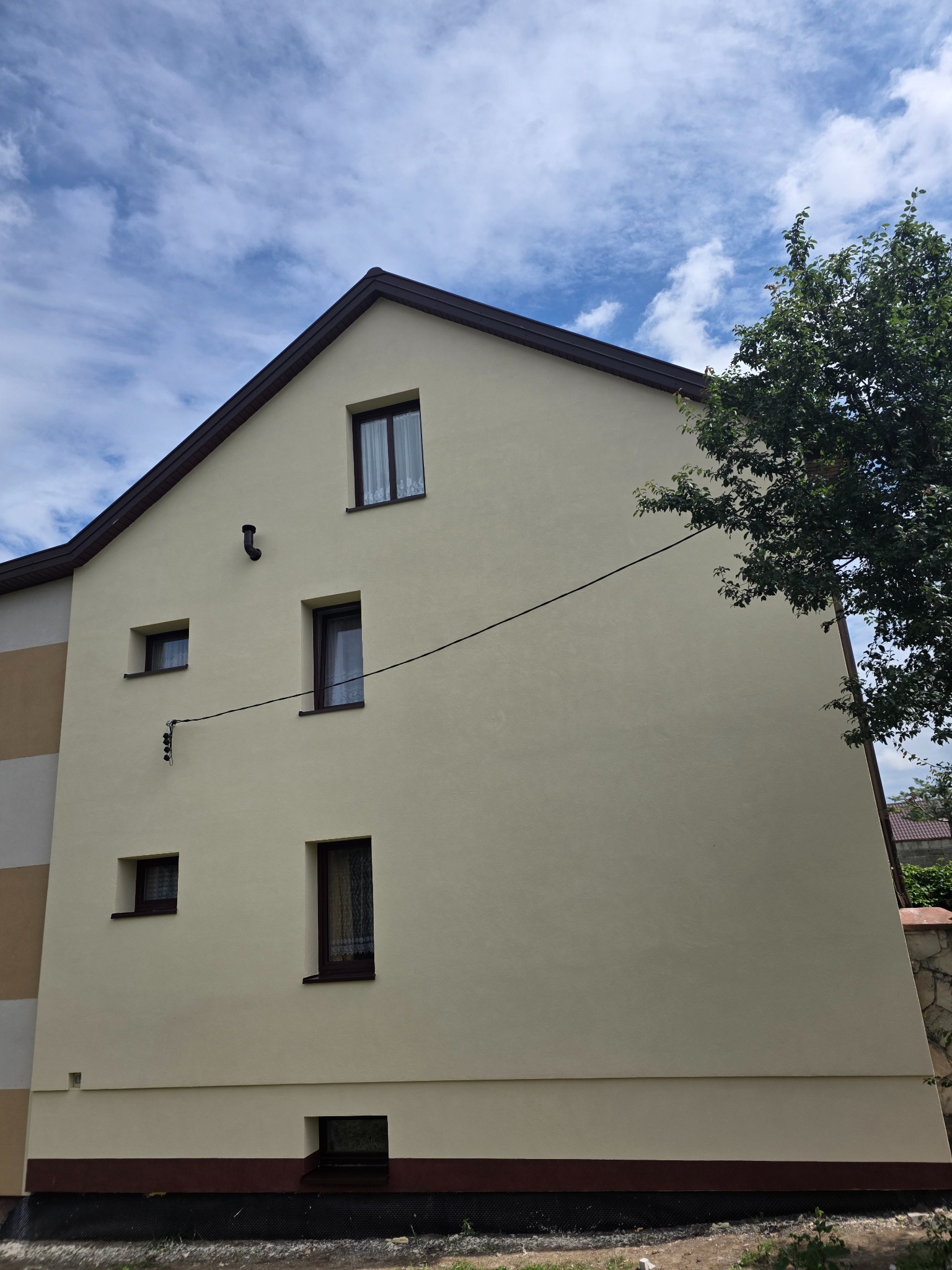 Elewacja domu po malowaniu w odcieniach beżu, z brązowymi ramami okiennymi i dachem. Widoczne okna o różnych rozmiarach i fragment drzewa po prawej stronie.