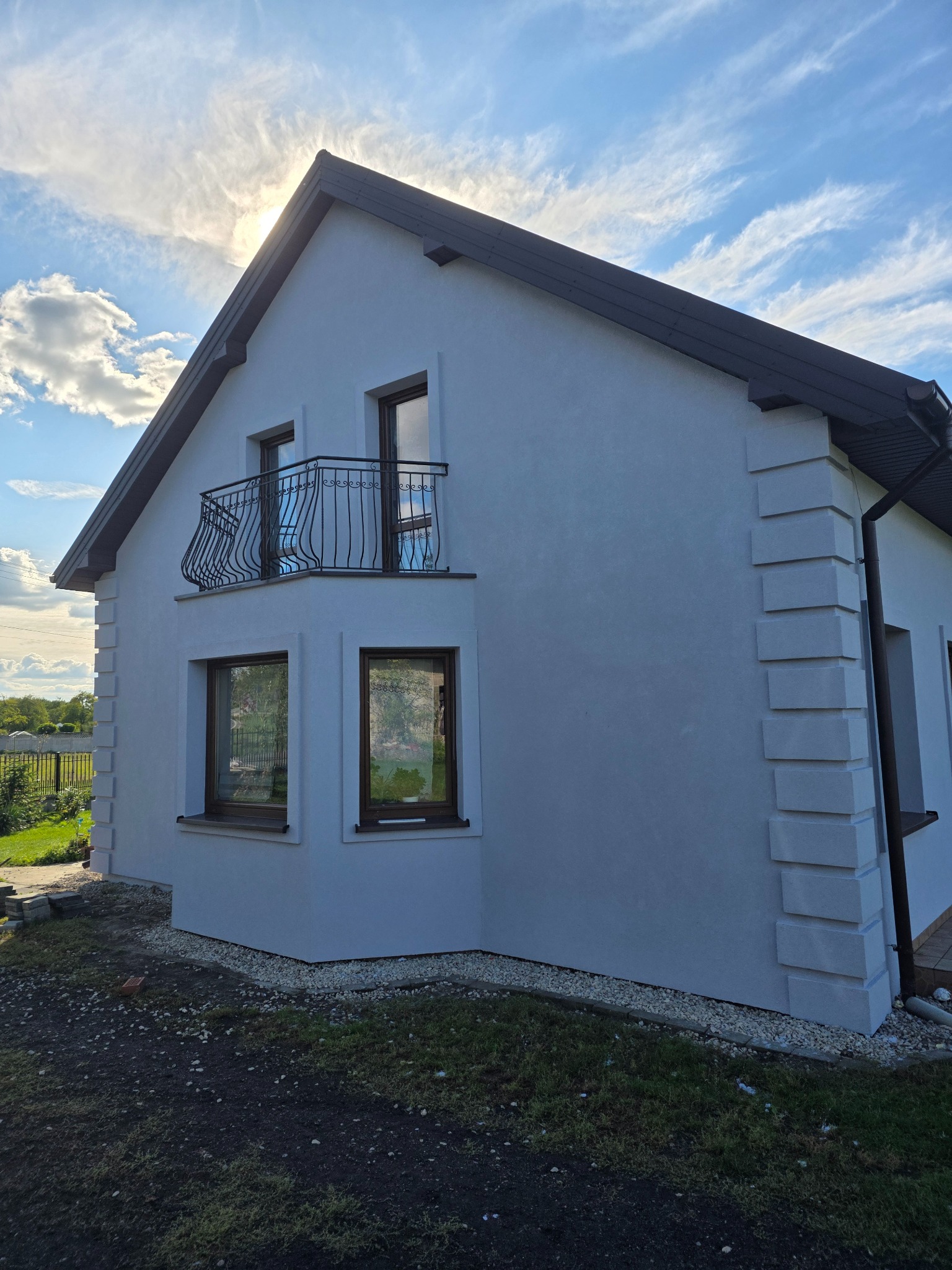 Biały dom z balkonem i dekoracyjnymi elementami elewacji w słoneczny dzień. Widoczne okna z brązowymi ramami i czarna rynna.