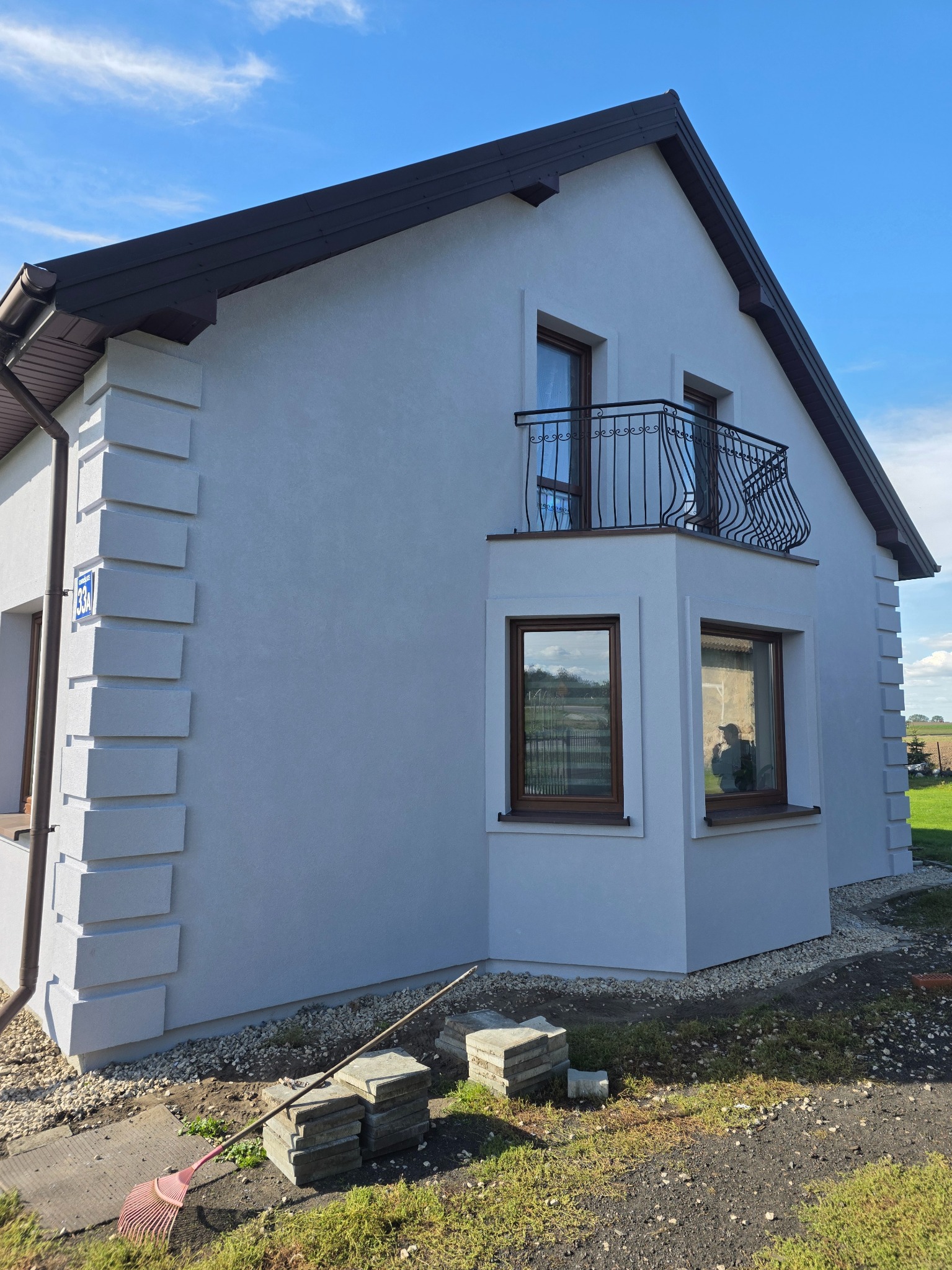 Elewacja domu jednorodzinnego po malowaniu w Orońsku. Jasny tynk, ciemne okna i dach. Widoczny balkon z ozdobną balustradą i dekoracyjne elementy na narożnikach.