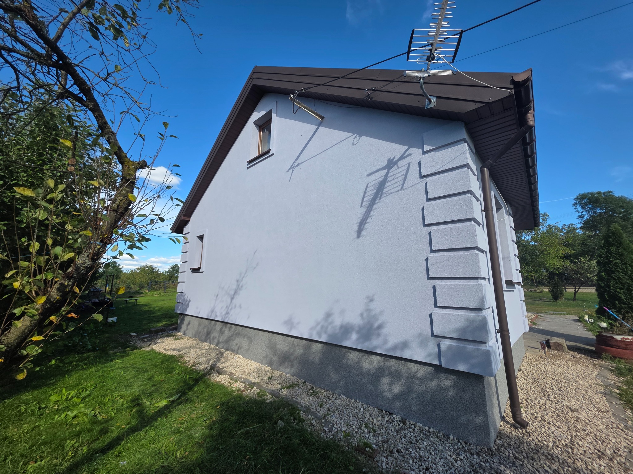 Świeżo pomalowana elewacja domu jednorodzinnego w odcieniach szarości, z dekoracyjnymi elementami architektonicznymi i brązowym dachem, na tle błękitnego nieba.