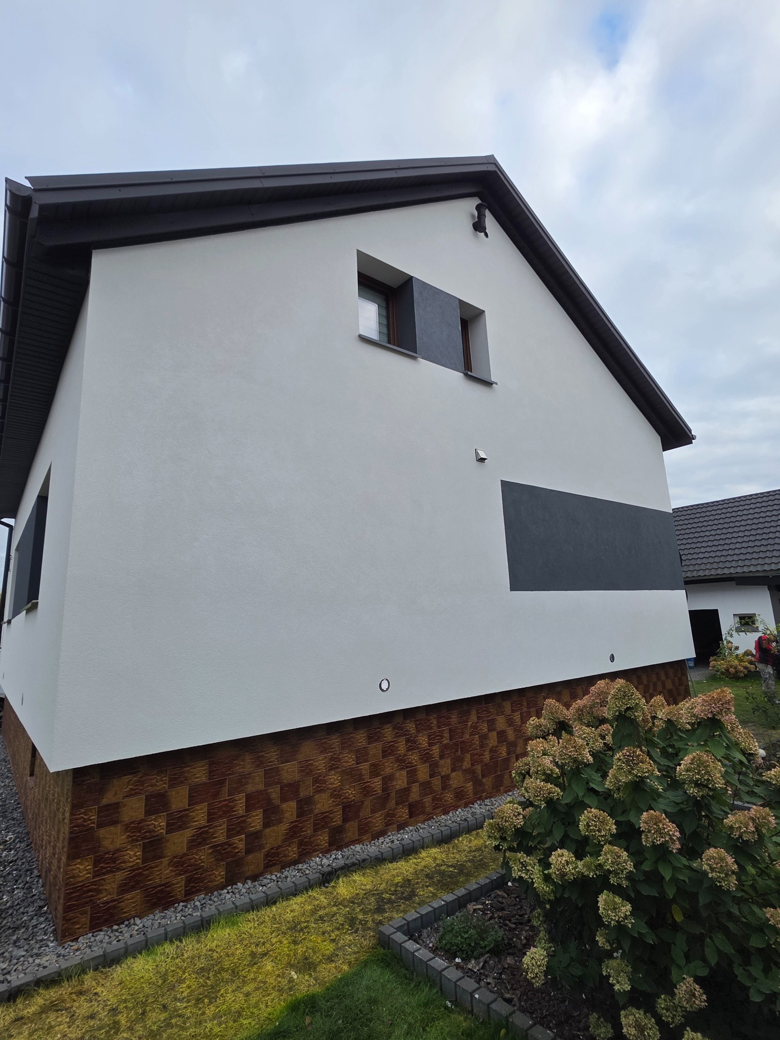 Ocieplony dom jednorodzinny z białą elewacją i brązowym cokołem, z szarym prostokątnym akcentem na ścianie. Widok z dołu, z krzewem w pierwszym planie.