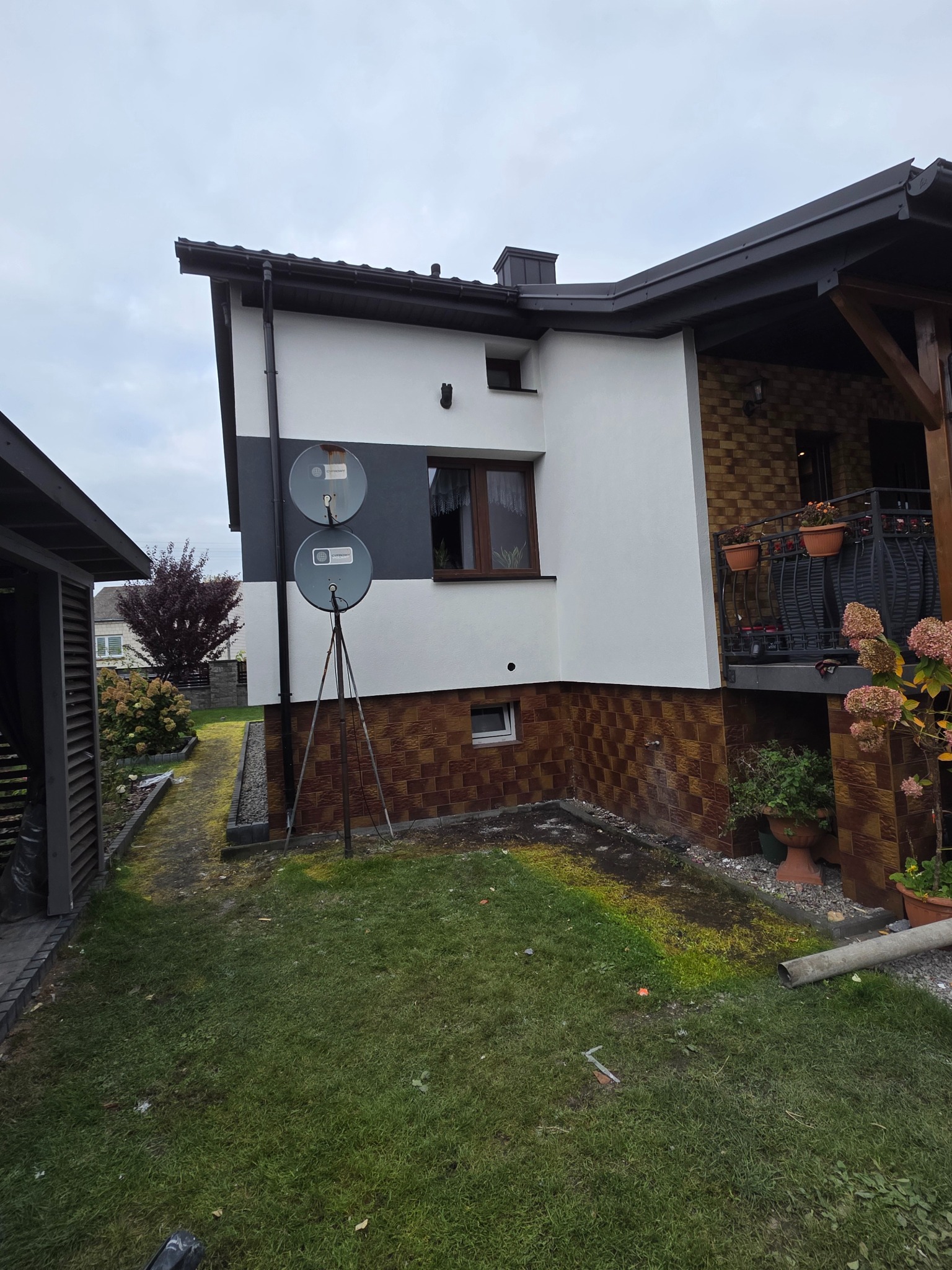 Dom jednorodzinny po renowacji elewacji: białe ściany z szarym pasem, brązowa cegła na dole, balkon z kwiatami, dwie anteny satelitarne na elewacji.