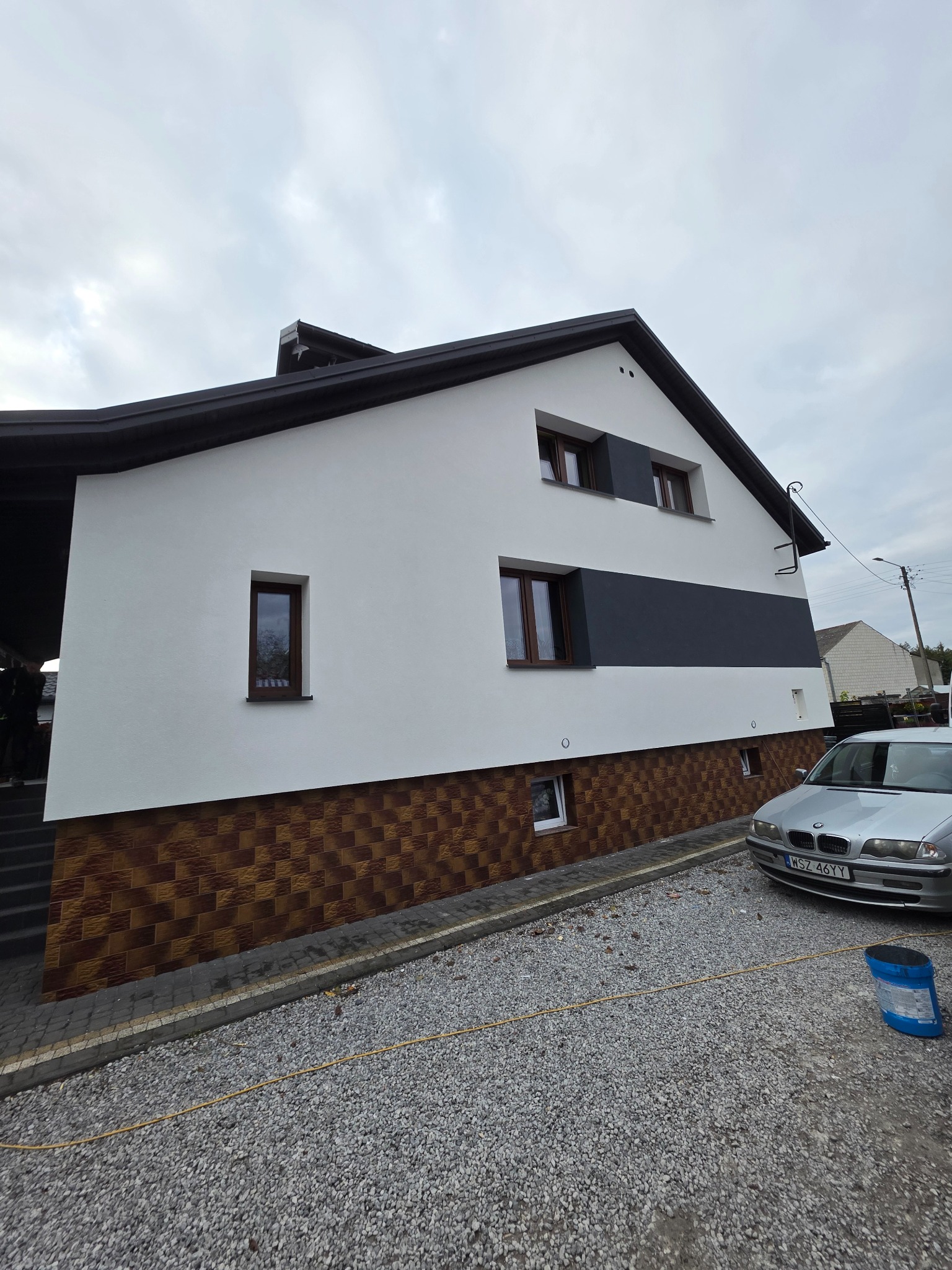 Dwukondygnacyjny dom z białą elewacją i brązową okładziną imitującą cegłę na dole. Widoczny szary pas na elewacji i zaparkowany samochód.