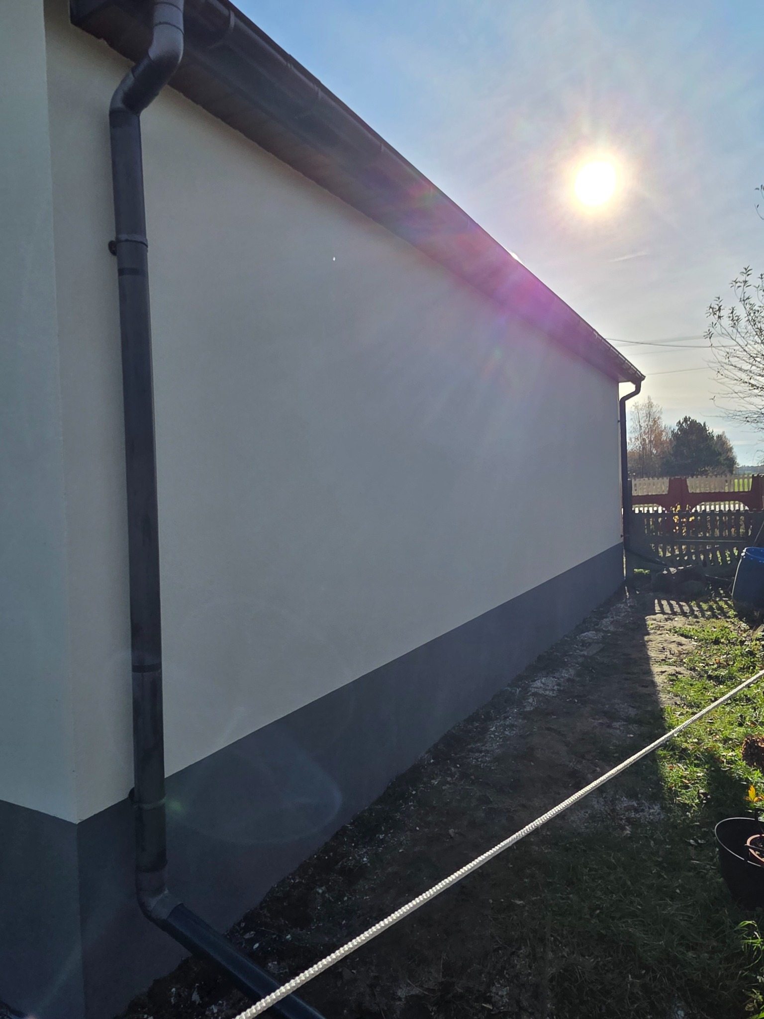 Ściana budynku po malowaniu elewacji w odcieniach szarości i bieli, z widocznym systemem rynnowym i fragmentem ogrodu w słoneczny dzień.