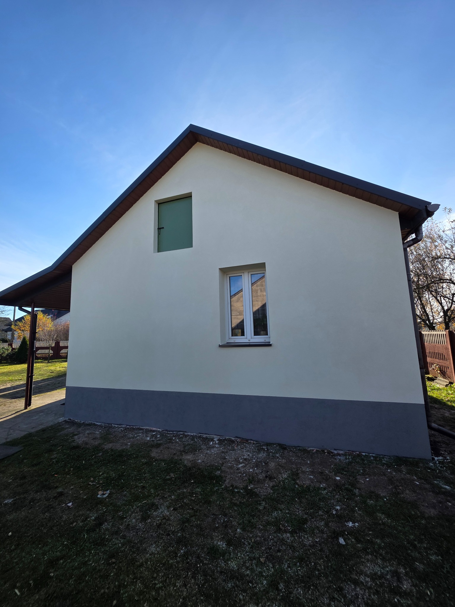 Jasna elewacja domu jednorodzinnego z szarą podmurówką i brązowym dachem w słoneczny dzień. Widoczne okno i małe okienko wentylacyjne. Trawa na pierwszym planie.