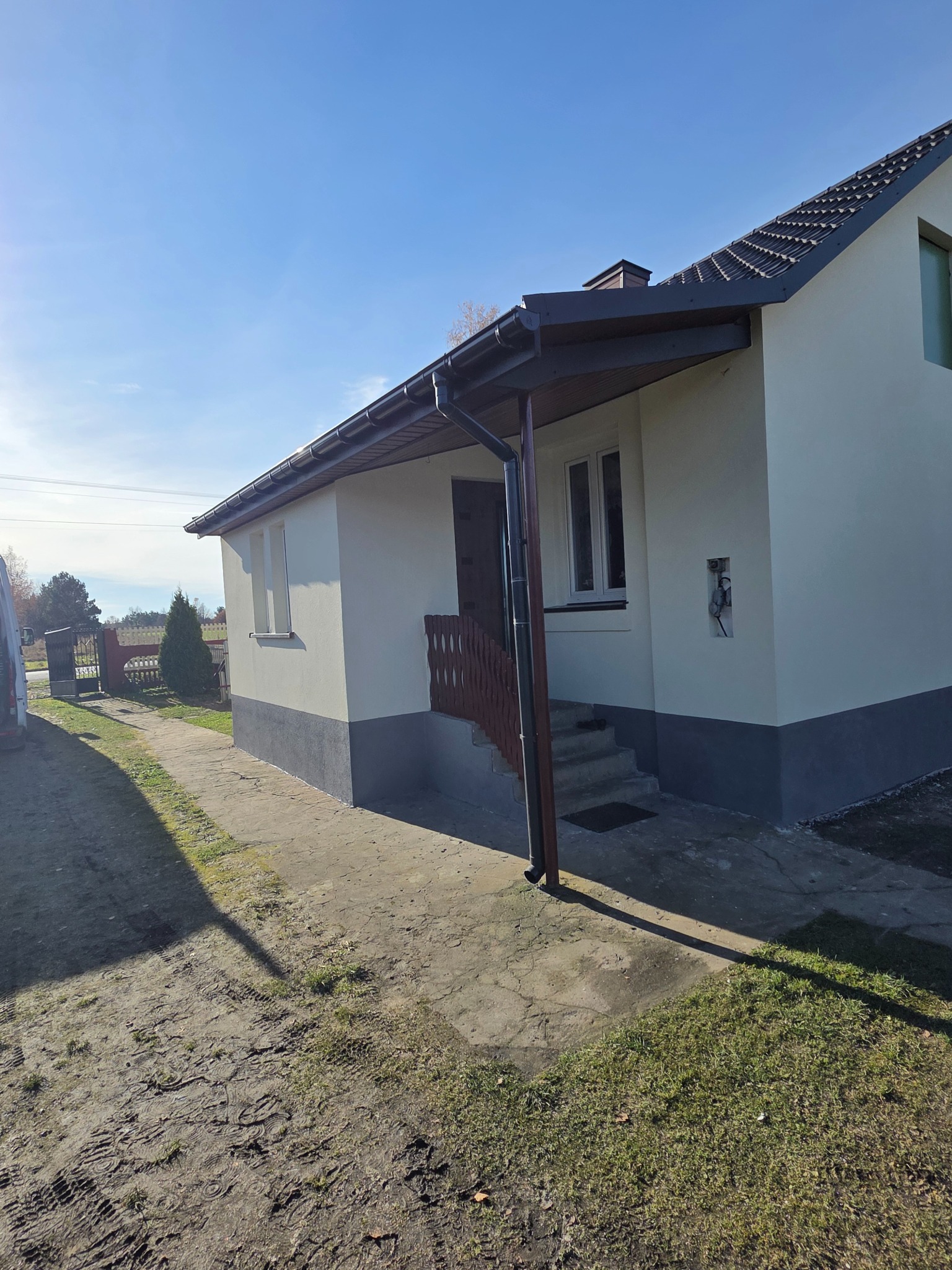 Jasny dom jednorodzinny z ciemną podmurówką i brązowymi rynnami w słoneczny dzień. Widoczne schody wejściowe i fragment ogrodu.