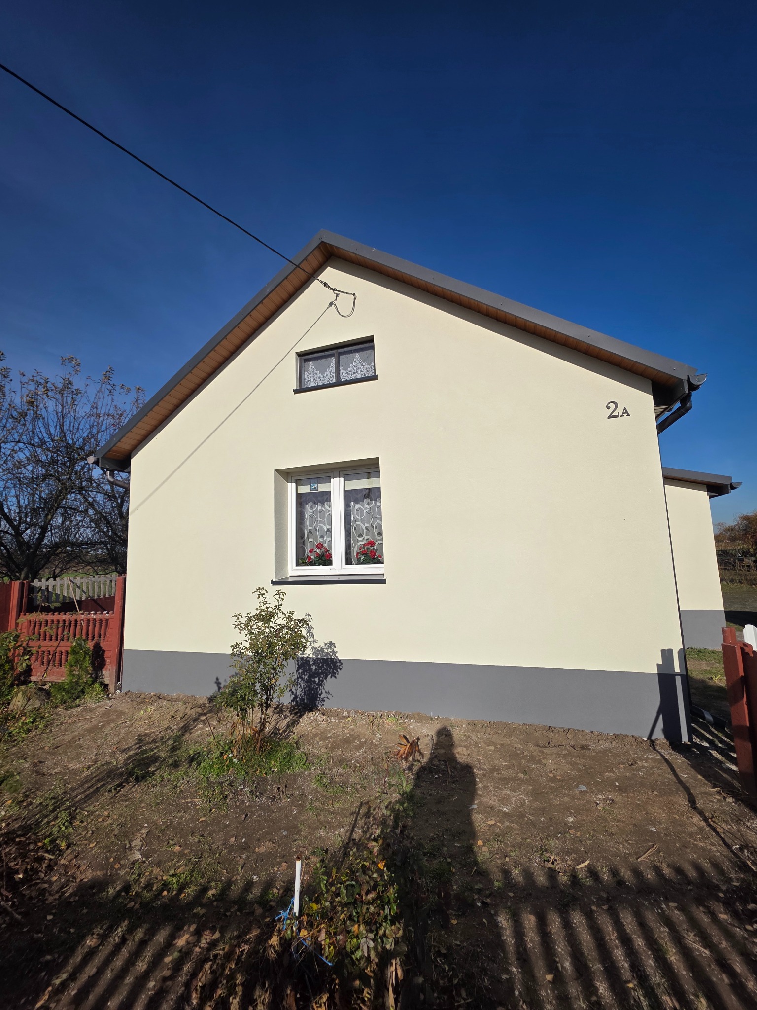 Jasna elewacja domu jednorodzinnego z numerem '2a' w słoneczny dzień. Widoczne okna z kwiatami i fragment czerwonego ogrodzenia. Czyste niebo w tle.