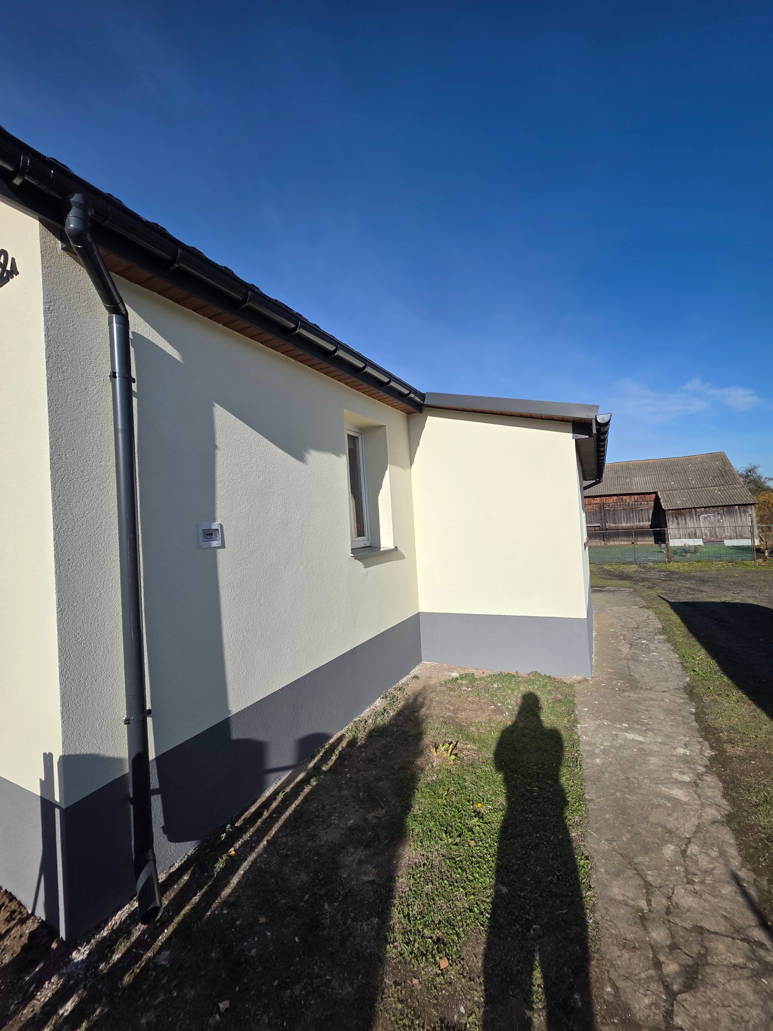 Ocieplony budynek z kremową elewacją i szarym cokołem, widoczny fragment rynny, okno i cień osoby na chodniku. W tle budynek gospodarczy i błękitne niebo.
