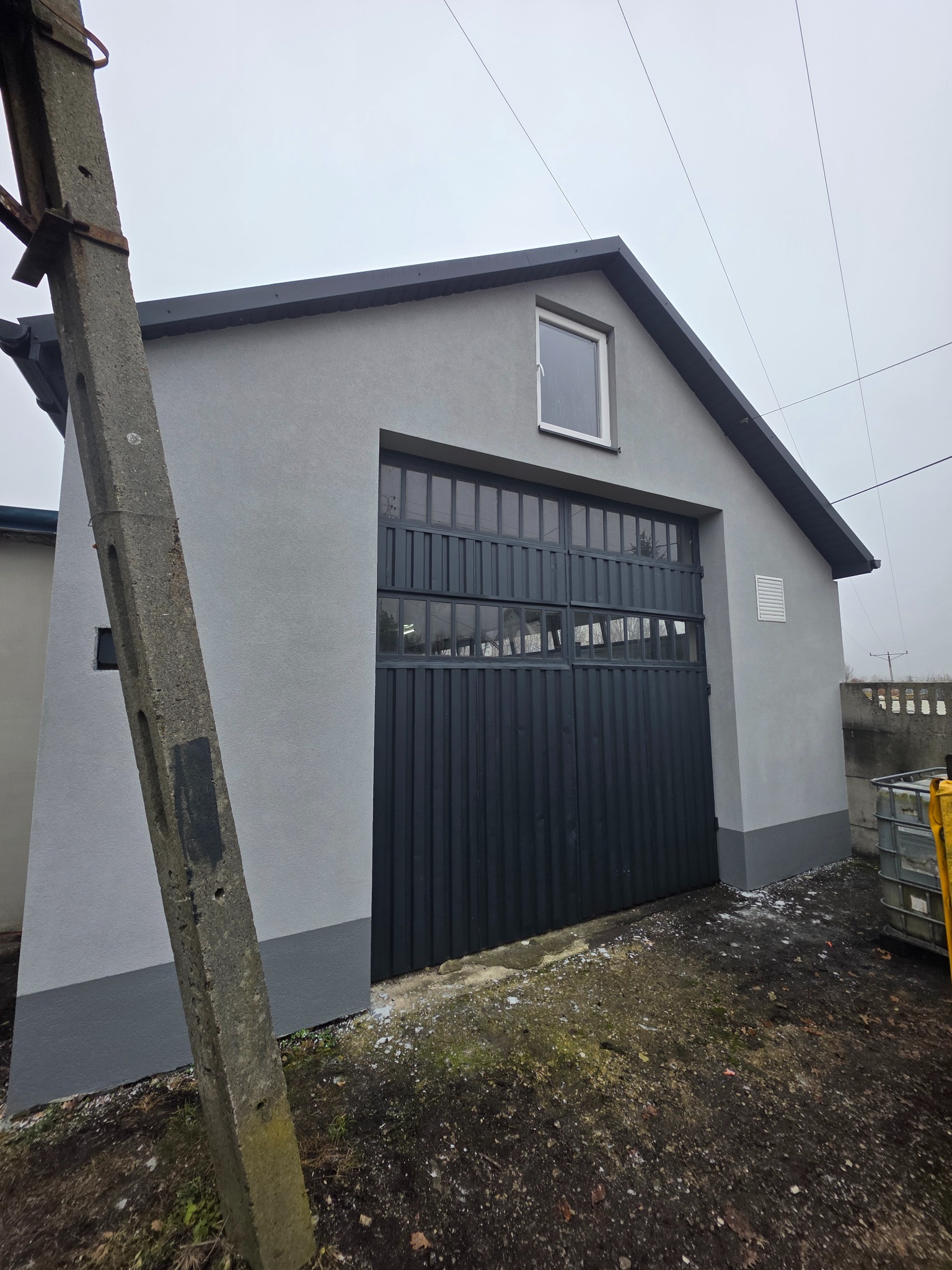Szary budynek gospodarczy z czarną bramą garażową, po świeżym malowaniu elewacji. Widoczny słup energetyczny i fragment otoczenia. Ujęcie z dołu, pochmurny dzień.