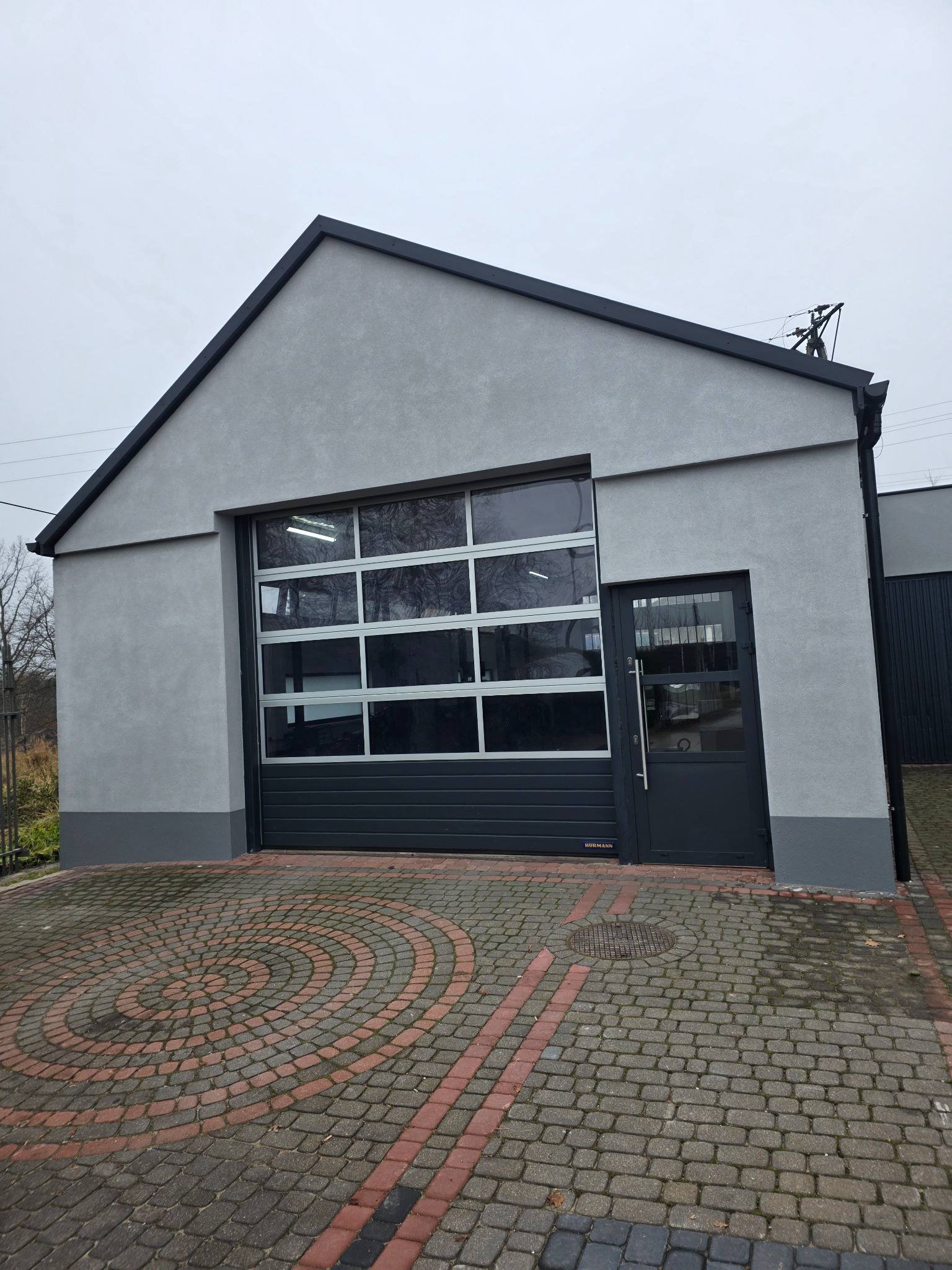 Szary budynek z bramą garażową segmentową i drzwiami wejściowymi, wykończony w nowoczesnym stylu. Kostka brukowa przed budynkiem.