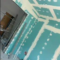 Ściana z płyt gipsowo-kartonowych w trakcie szpachlowania, widoczne łączenia i śruby, fragment sufitu z konstrukcją metalową, czerwona skrzynka narzędziowa Hilti na podłodze.