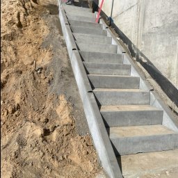 Paweł Pyrek - Wykonane betonowe schody zewnętrzne prowadzące z nasypu ziemnego do betonowego muru, widoczne buty robotnika na górnym stopniu i fragment drugiego robotnika w niebieskim kombinezonie na nasypie.