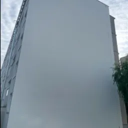 Pomalowana na szaro ściana bloku mieszkalnego, widziana z dołu, z widocznym zielonym śmietnikiem i małą choinką u podstawy ściany.