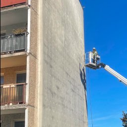 Paweł Pyrek - Pracownik w podnośniku koszowym wykonuje prace na ocieplonej ścianie bloku mieszkalnego, widoczne ślady po kołkach mocujących styropian, balkony z metalowymi barierkami i roślinami.