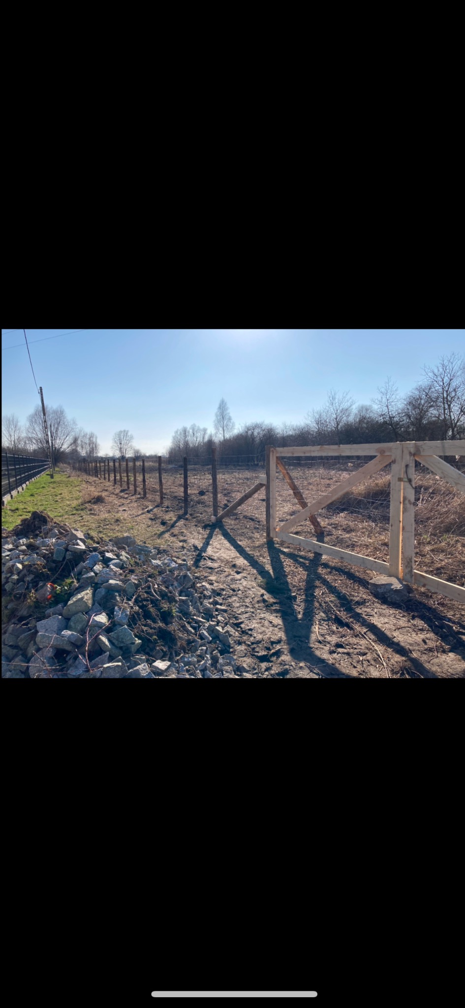 Nowo postawione drewniane ogrodzenie z furtką na nierównym terenie, widoczne kamienie i słupki w perspektywie, w tle drzewa bez liści pod błękitnym niebem.