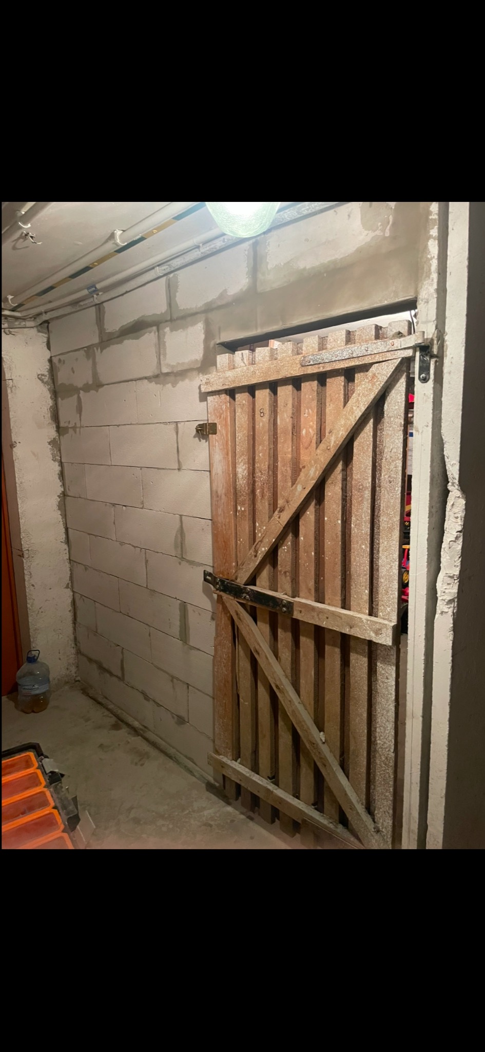 Surowa drewniana brama z ukośnymi wzmocnieniami w piwnicy z niedokończonymi ścianami z bloczków betonowych i widocznymi rurami pod sufitem.