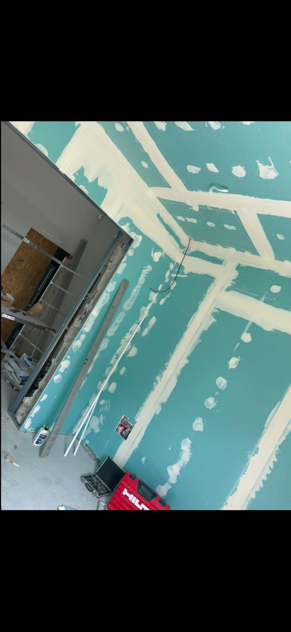 Ściana z płyt gipsowo-kartonowych w trakcie szpachlowania, widoczne łączenia i śruby, fragment sufitu z konstrukcją metalową, czerwona skrzynka narzędziowa Hilti na podłodze.