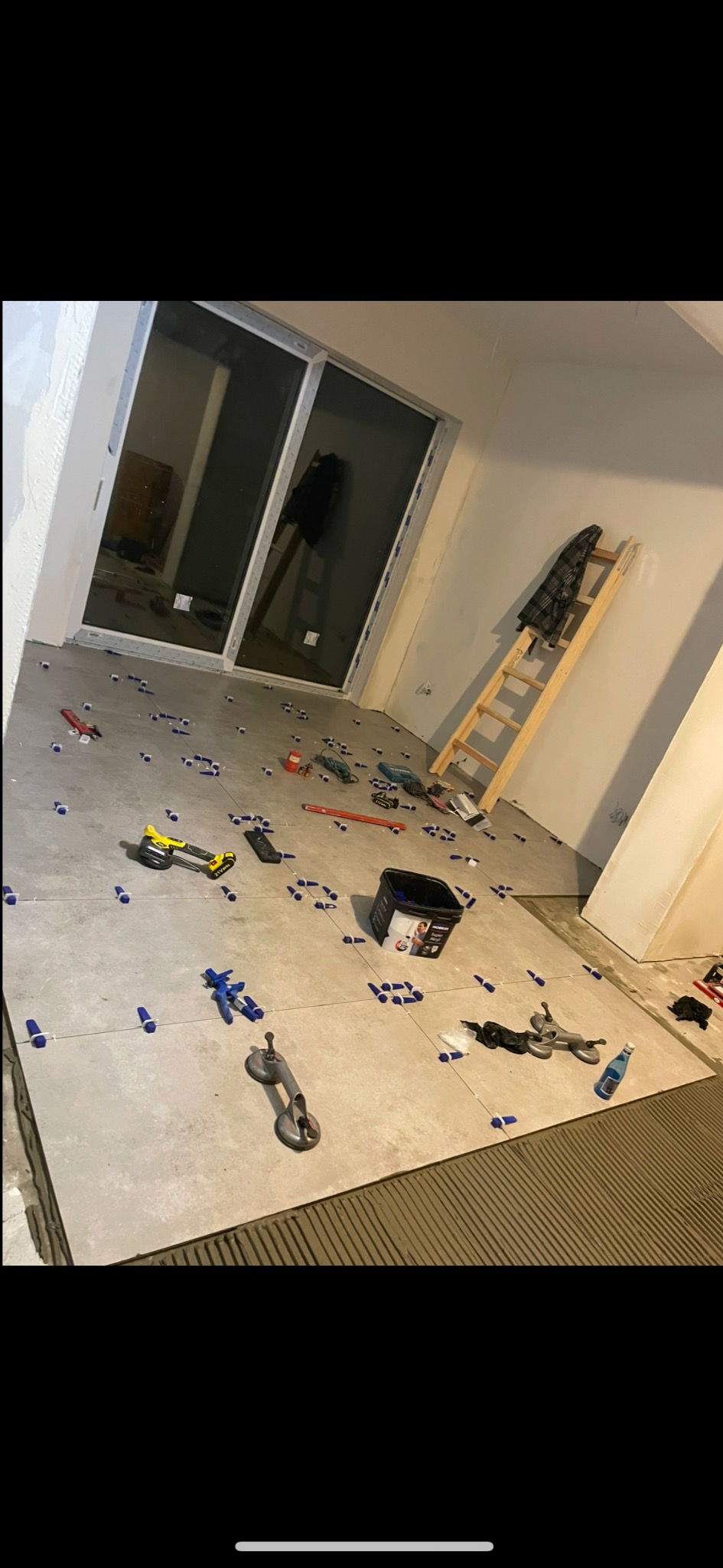 Układanie płytek podłogowych w pomieszczeniu z dużym oknem balkonowym, widoczne narzędzia glazurnicze, poziomica, szpachelka, przyssawki do płytek oraz drabina oparta o ścianę.