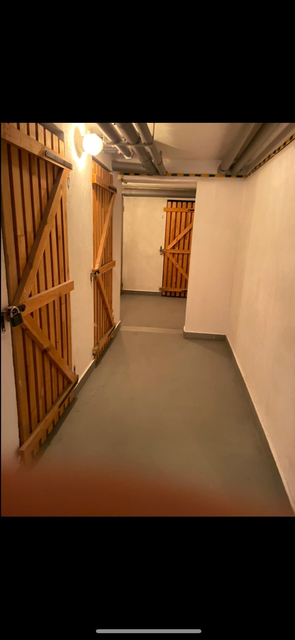 Korytarz piwniczny z drewnianymi drzwiami do komórek lokatorskich, szara posadzka z wylewki, rury pod sufitem z żółto-czarnym pasem ostrzegawczym.