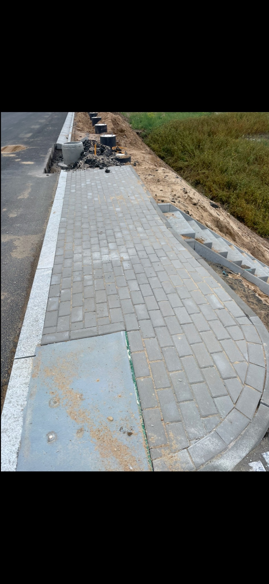 Ułożona szara kostka brukowa tworząca ścieżkę, z widocznymi elementami kanalizacji i przygotowanym terenem pod dalsze prace ziemne.