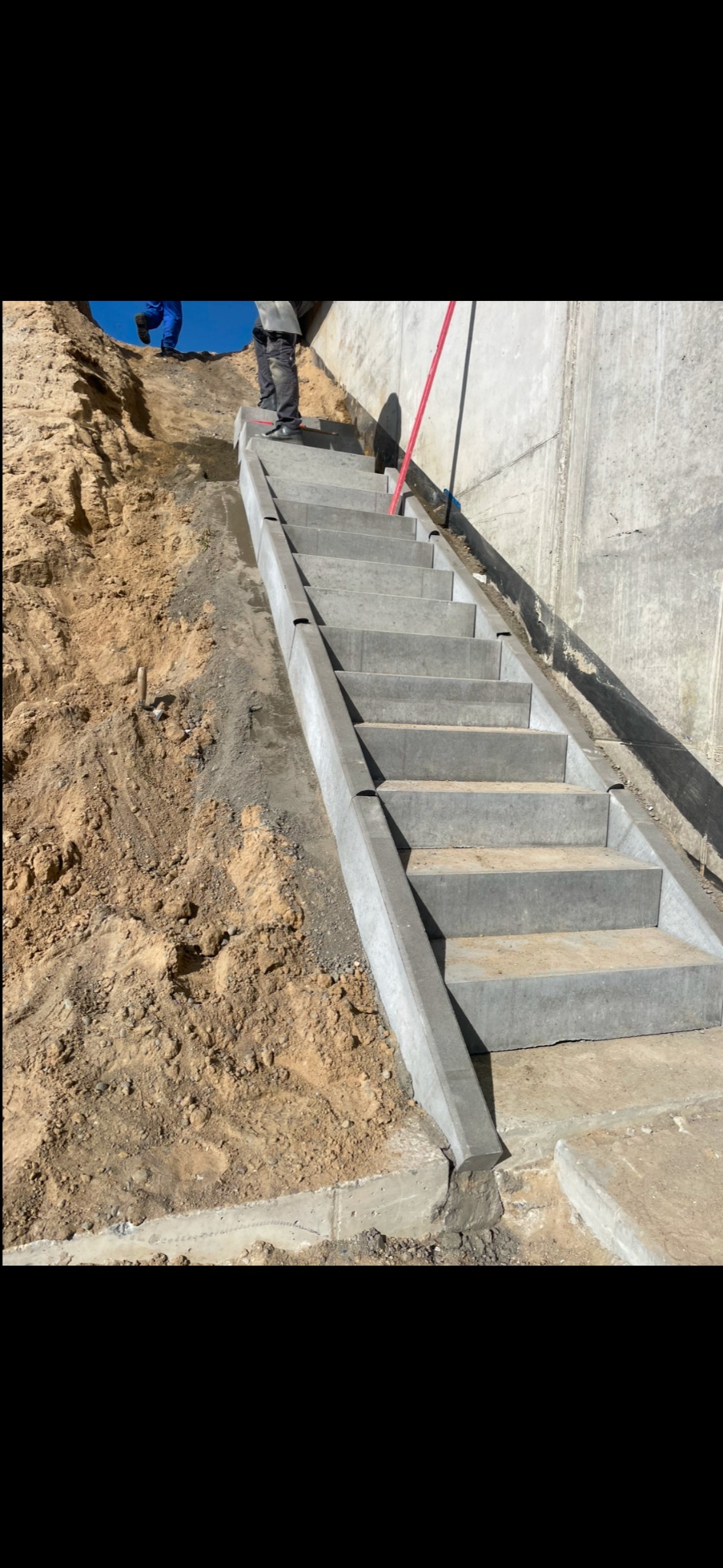 Wykonane betonowe schody zewnętrzne prowadzące z nasypu ziemnego do betonowego muru, widoczne buty robotnika na górnym stopniu i fragment drugiego robotnika w niebieskim kombinezonie na nasypie.