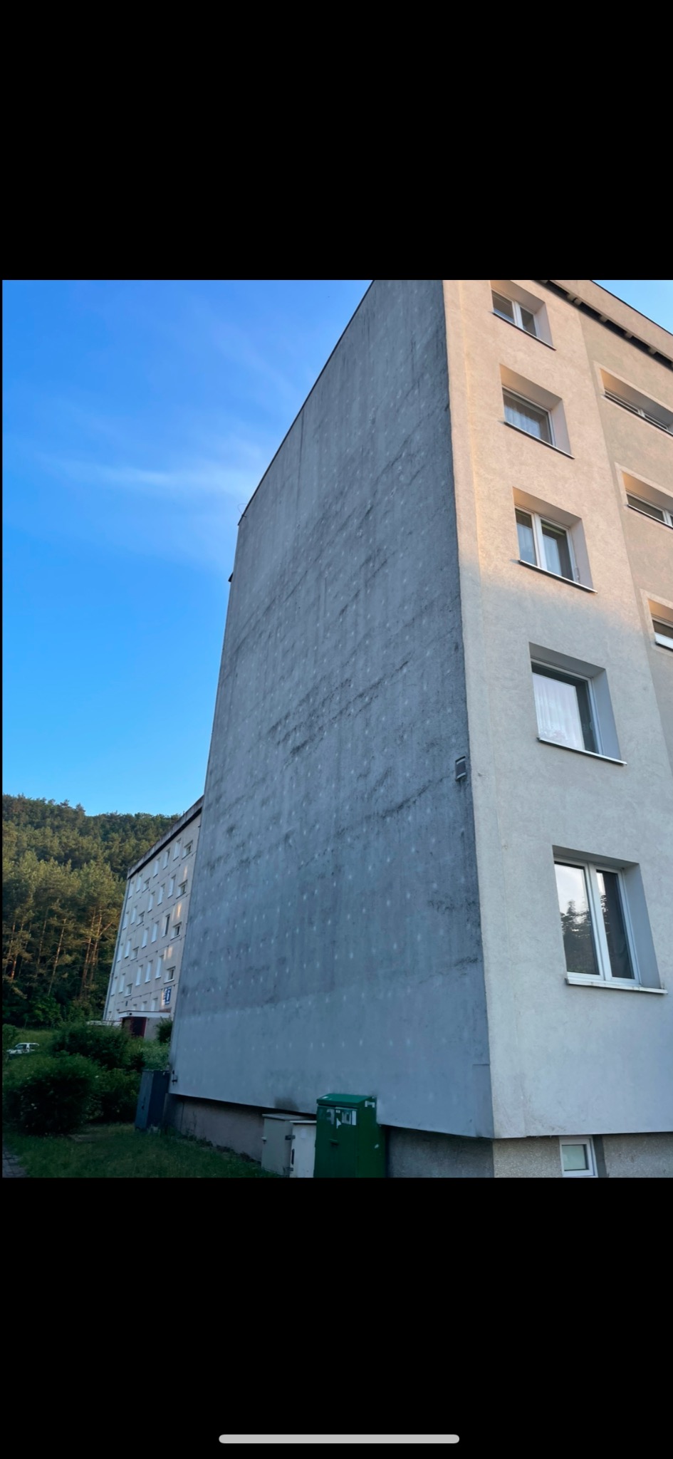 Szary blok mieszkalny z widocznymi śladami po zaciekach na ścianie, zieleń drzew w tle, ujęcie z dołu pod kątem.