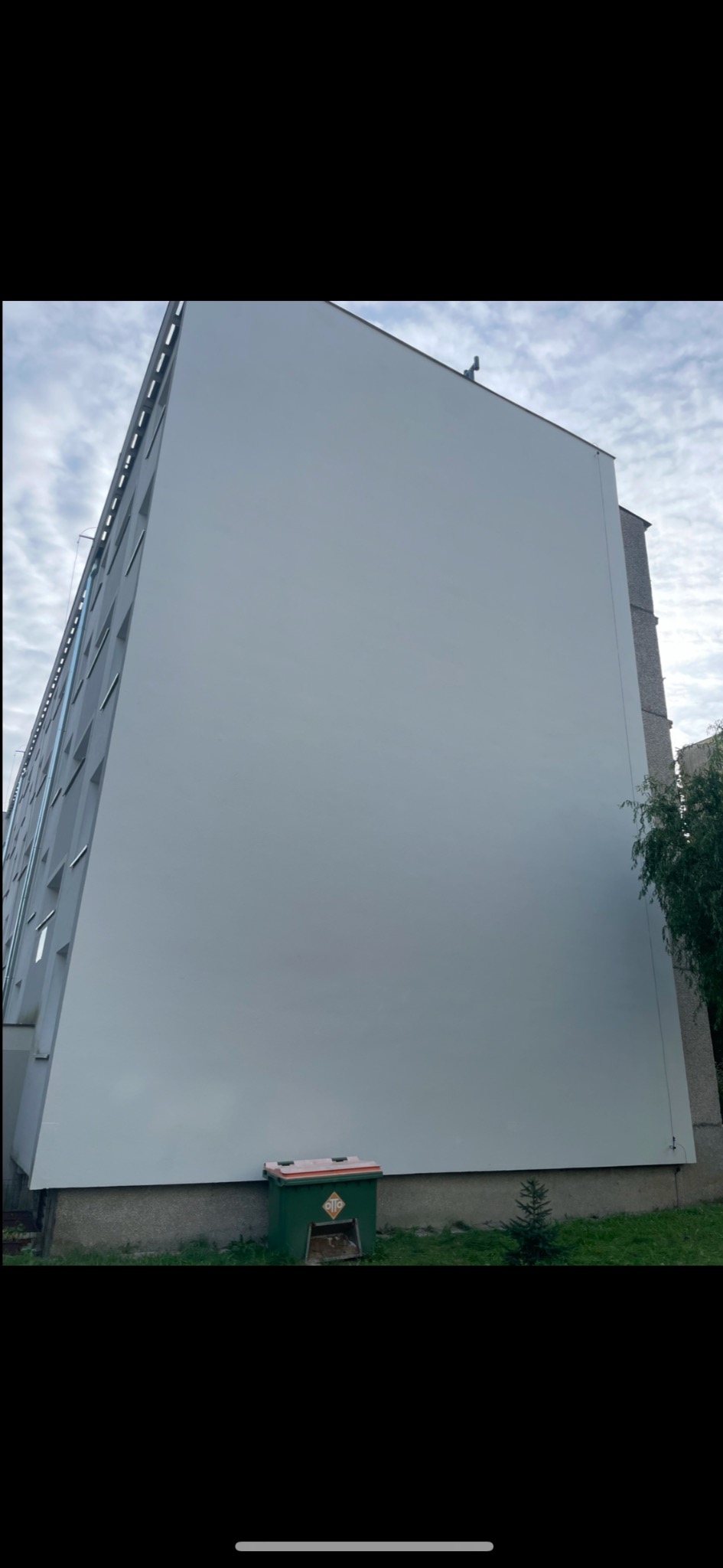 Pomalowana na szaro ściana bloku mieszkalnego, widziana z dołu, z widocznym zielonym śmietnikiem i małą choinką u podstawy ściany.