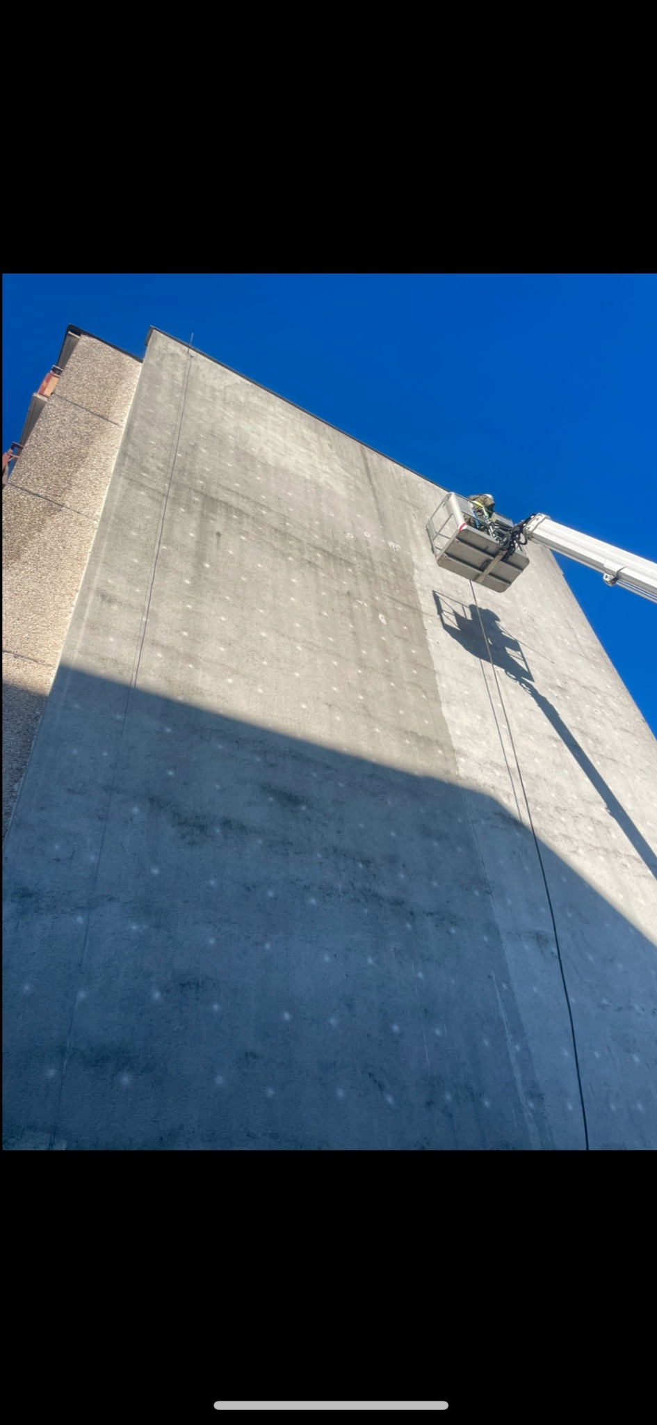 Pracownik w podnośniku koszowym myje elewację wysokiego budynku, widoczne plamy na ścianie i ślady po myciu.