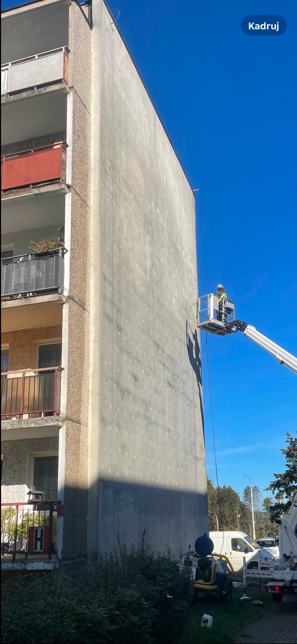 Pracownik w podnośniku koszowym wykonuje prace na ocieplonej ścianie bloku mieszkalnego, widoczne ślady po kołkach mocujących styropian, balkony z metalowymi barierkami i roślinami.