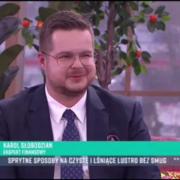 Mężczyzna w garniturze i okularach, Karol Słobodzian, ekspert finansowy, występuje w programie telewizyjnym na tle dekoracji roślinnych.