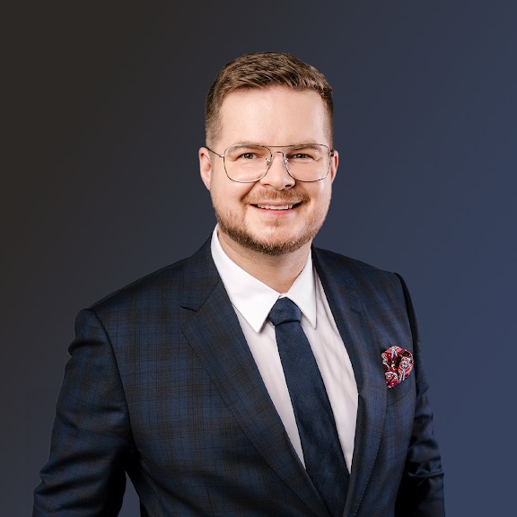 Portret uśmiechniętego mężczyzny w okularach, w granatowym garniturze w kratę i z bordową poszetką w kieszonce, na ciemnym tle.