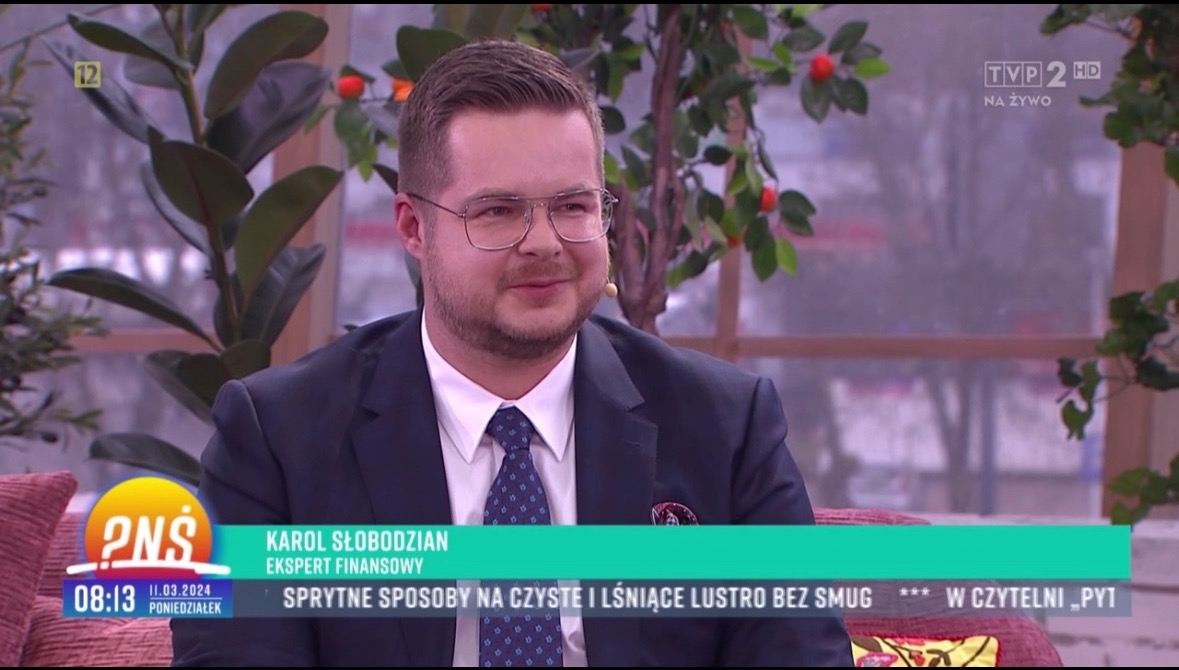 Mężczyzna w garniturze i okularach, Karol Słobodzian, ekspert finansowy, występuje w programie telewizyjnym na tle dekoracji roślinnych.