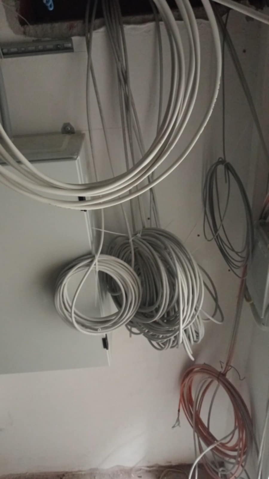Pętle białych i pomarańczowych kabli zwisające z sufitu podczas prac remontowych, widoczna skrzynka elektryczna i fragment listwy montażowej.
