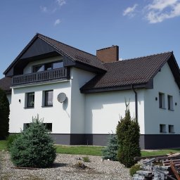 FIRMA HANDLOWO USŁUGOWA DAMIAN BALCAR - Nowoczesny dom z białą elewacją, ciemną stolarką okienną i dachem z brązowej dachówki, antena satelitarna na ścianie, w tle zielony trawnik i drzewa, na pierwszym planie niskie iglaki i stos...