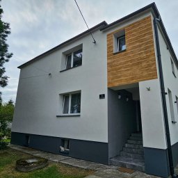 FIRMA HANDLOWO USŁUGOWA DAMIAN BALCAR - Dwukondygnacyjny dom z białą elewacją i drewnopodobną wstawką, ciemnoszara podmurówka, kot w oknie piwnicznym.