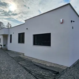 Nowoczesny, parterowy dom z płaskim dachem, wykończony jasnym tynkiem, z minimalistycznymi oknami z żaluzjami zewnętrznymi, oświetlony zachodzącym słońcem, z widoczną instalacją alarmową...