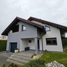 FIRMA HANDLOWO USŁUGOWA DAMIAN BALCAR - Dwukondygnacyjny dom z szarą i białą elewacją, brązowym dachem i oknami z roletami, z widocznymi schodami wejściowymi z kostki brukowej i stertą kamieni na trawniku.