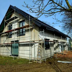 Dom jednorodzinny w trakcie tynkowania elewacji, widoczne rusztowania, drewniana drabina oparta o ścianę, świeżo posiana trawa wokół budynku, błękitne niebo w tle.