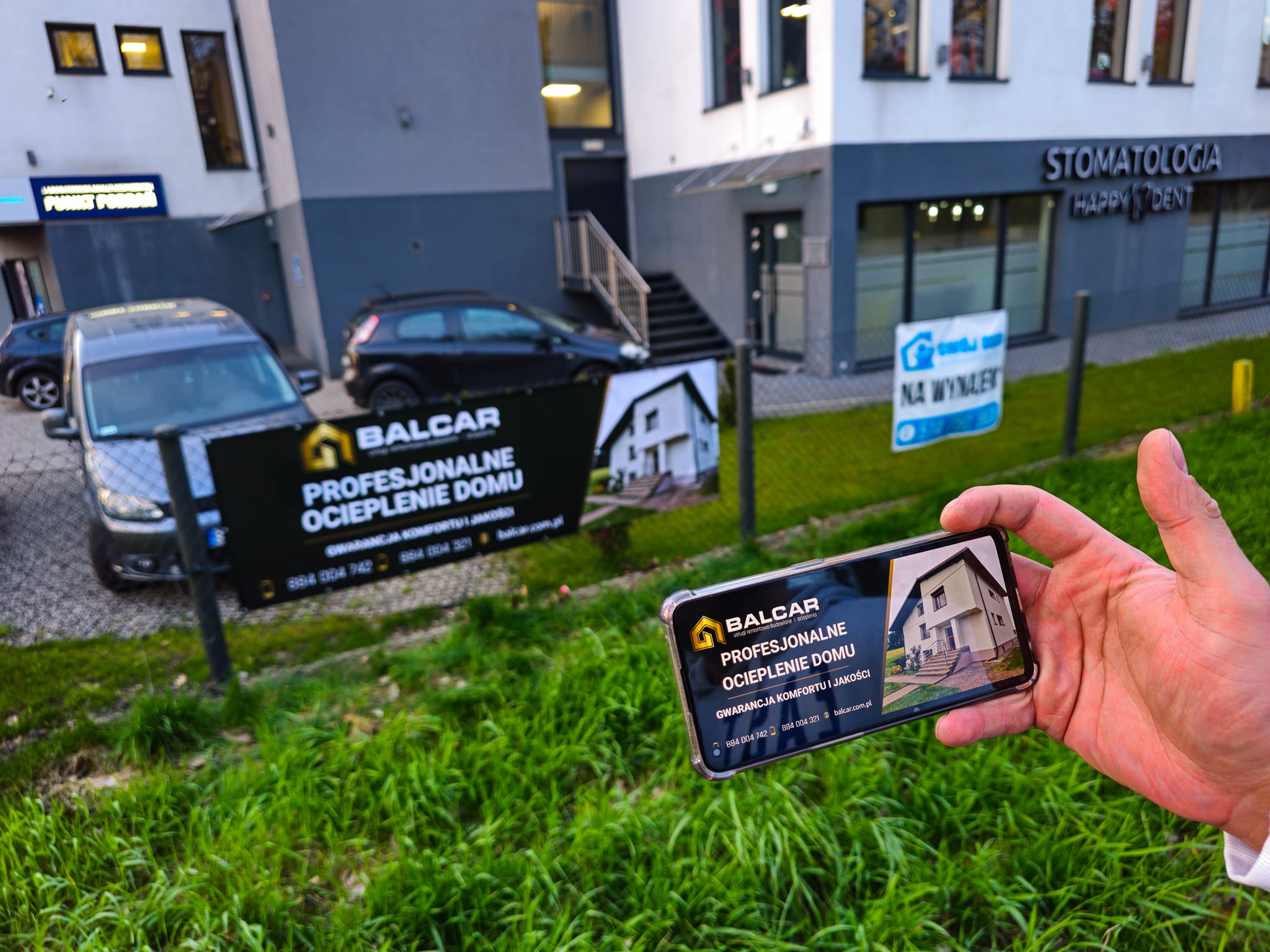 Zdjęcie prezentuje baner reklamowy firmy Balcar, specjalizującej się w profesjonalnym ociepleniu domu, widziany przez ekran smartfona trzymanego w dłoni, z budynkiem w tle i zieloną trawą...