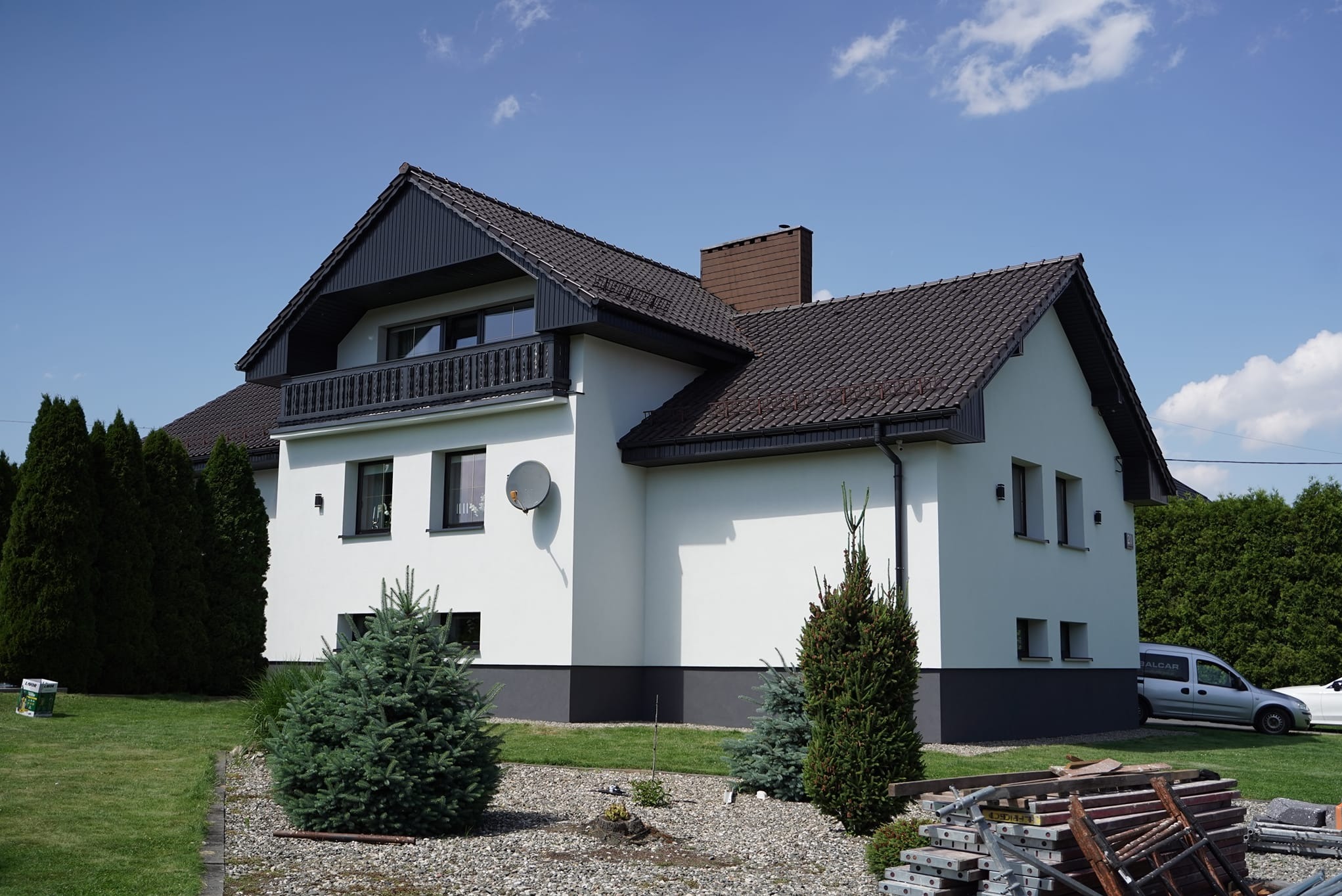 Nowoczesny dom z białą elewacją, ciemną stolarką okienną i dachem z brązowej dachówki, antena satelitarna na ścianie, w tle zielony trawnik i drzewa, na pierwszym planie niskie iglaki i stos...