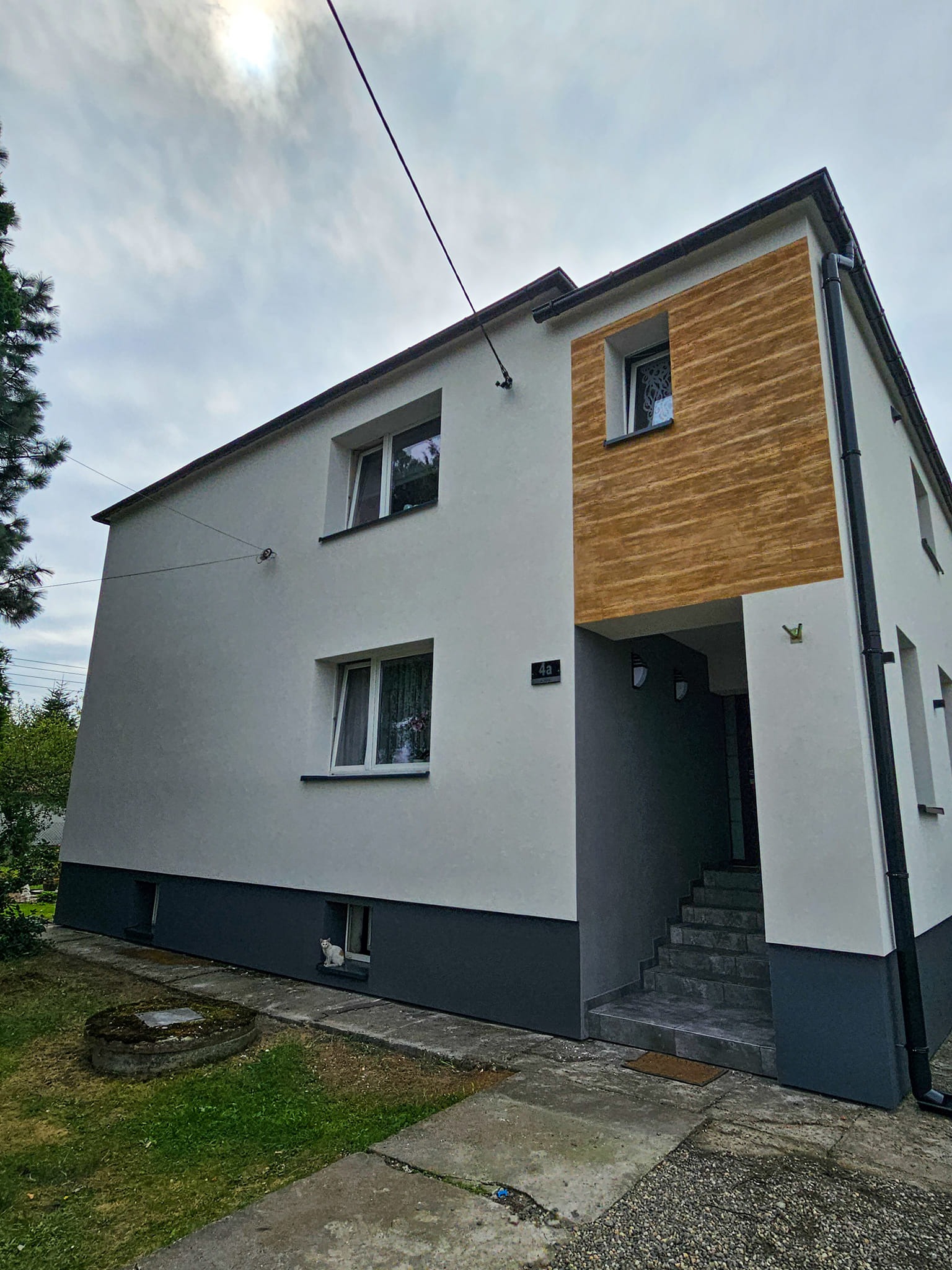 Dwukondygnacyjny dom z białą elewacją i drewnopodobną wstawką, ciemnoszara podmurówka, kot w oknie piwnicznym.