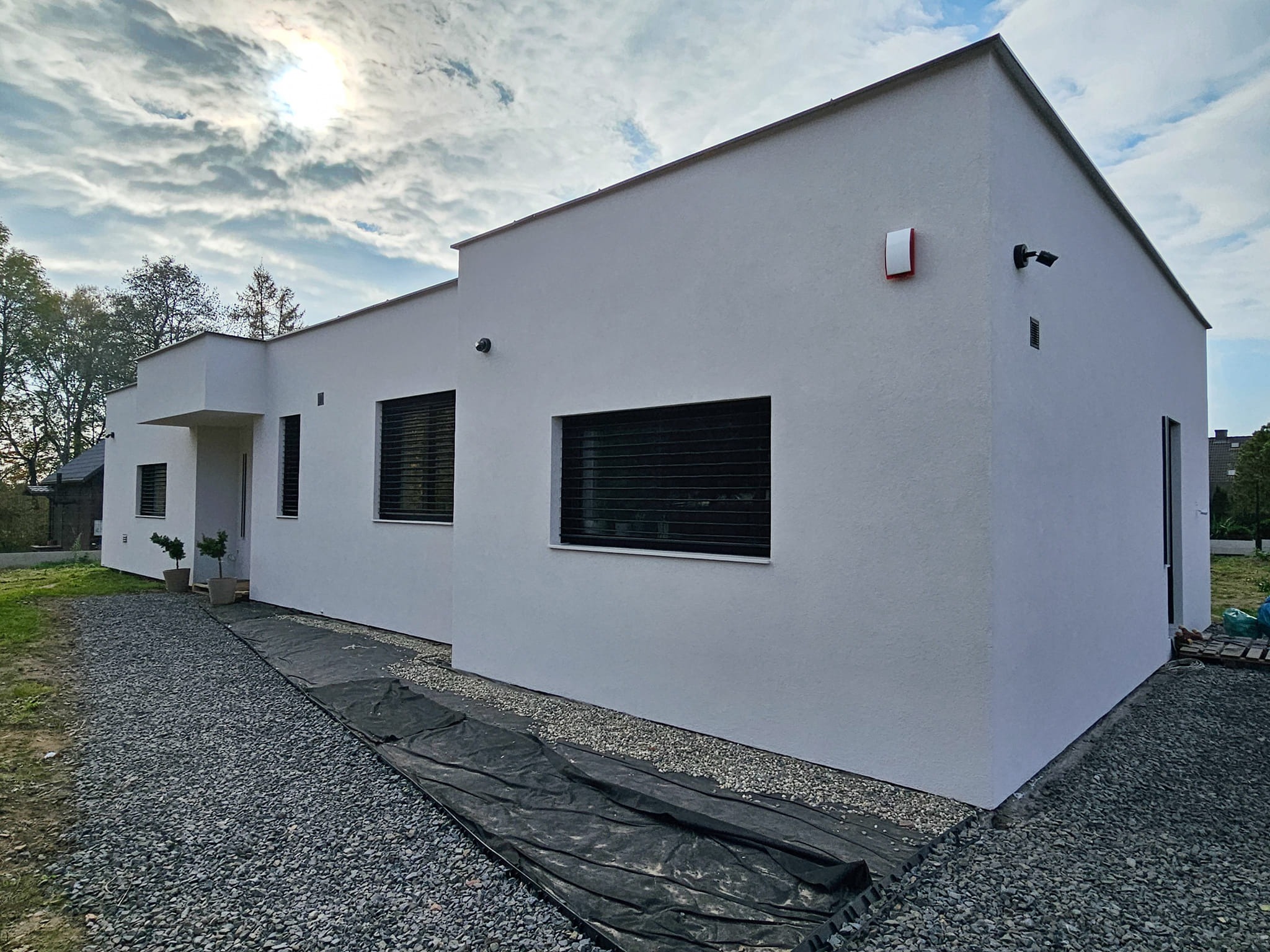 Nowoczesny, parterowy dom z płaskim dachem, wykończony jasnym tynkiem, z minimalistycznymi oknami z żaluzjami zewnętrznymi, oświetlony zachodzącym słońcem, z widoczną instalacją alarmową...