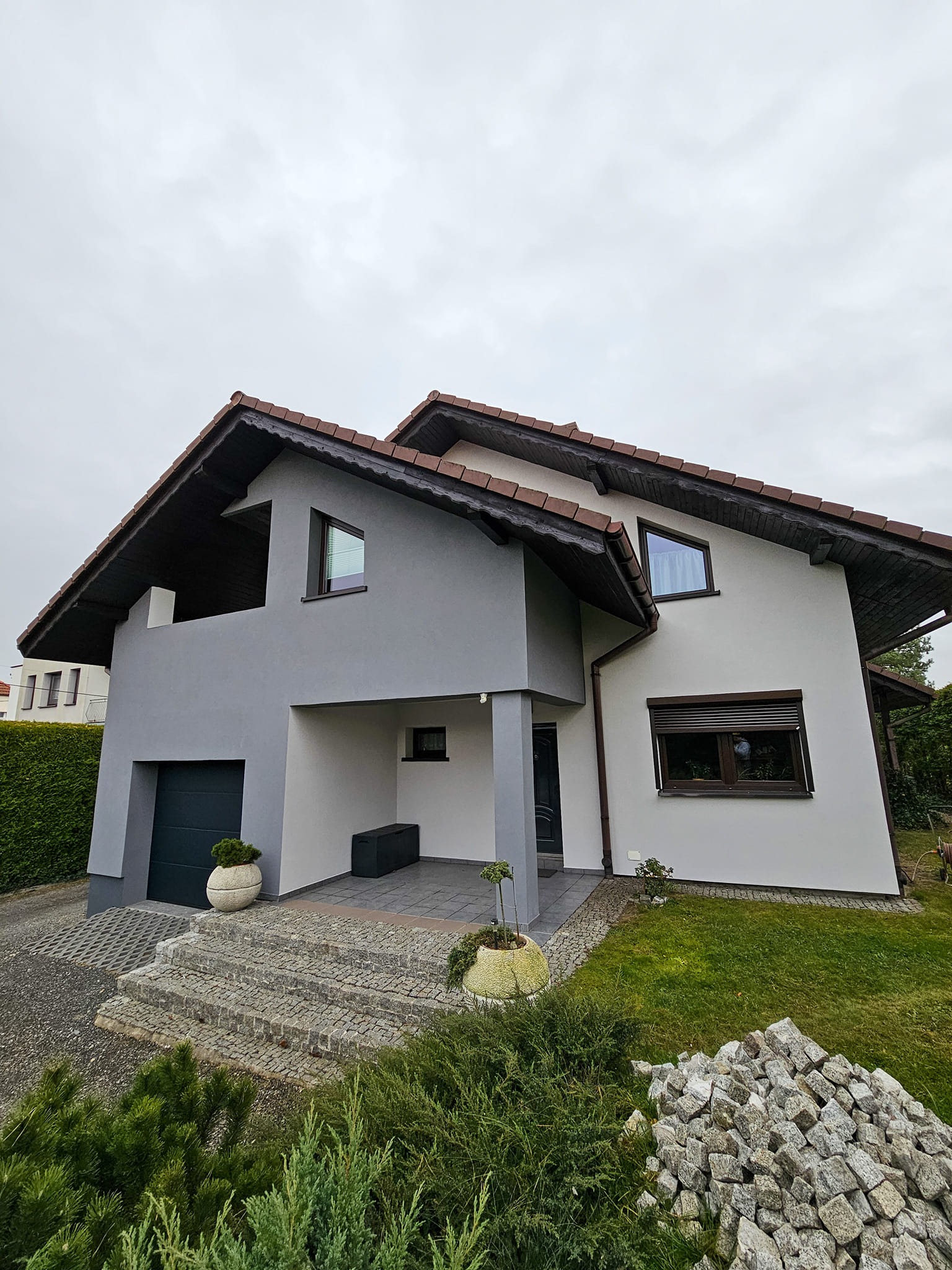 Dwukondygnacyjny dom z szarą i białą elewacją, brązowym dachem i oknami z roletami, z widocznymi schodami wejściowymi z kostki brukowej i stertą kamieni na trawniku.