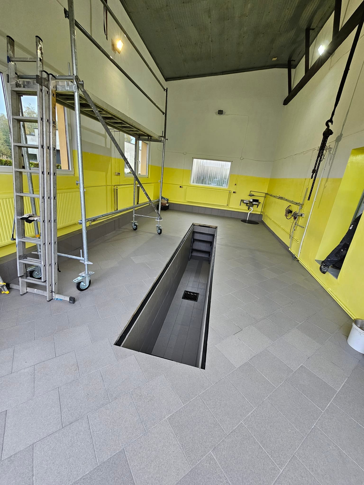 Wnętrze warsztatu samochodowego w trakcie remontu, widoczne szare płytki podłogowe, żółto-białe ściany, kanał naprawczy i rusztowanie na kółkach.