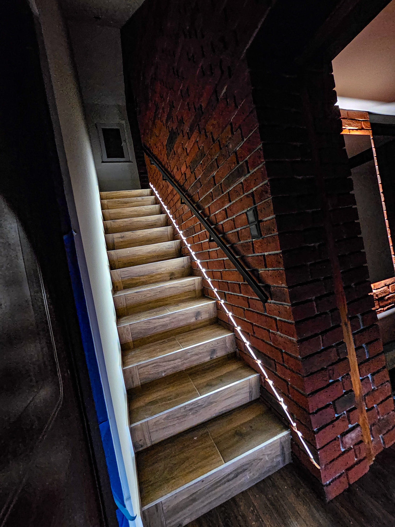 Schody z drewnopodobnych płytek z podświetleniem LED wzdłuż poręczy, ściana z czerwonej cegły, widok z dołu.