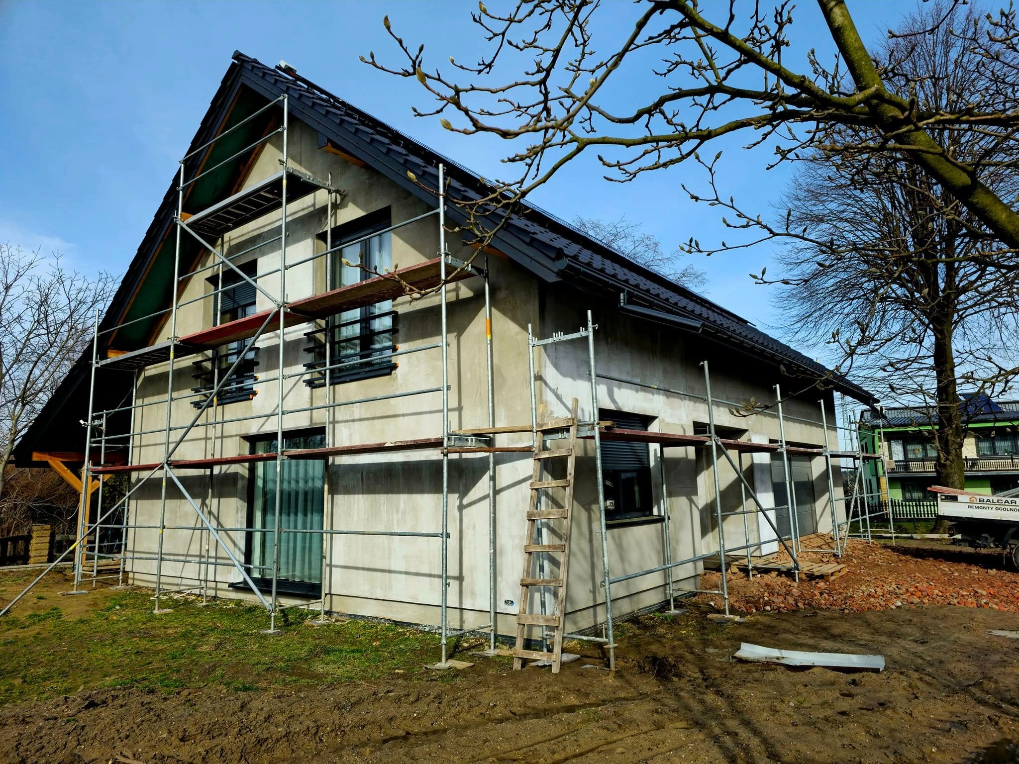 Dom jednorodzinny w trakcie tynkowania elewacji, widoczne rusztowania, drewniana drabina oparta o ścianę, świeżo posiana trawa wokół budynku, błękitne niebo w tle.