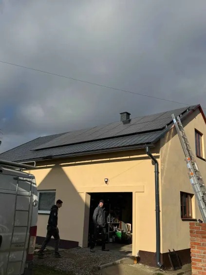 Montaż paneli słonecznych na dachu garażu z dwoma pracownikami i drabiną opartą o ścianę budynku.