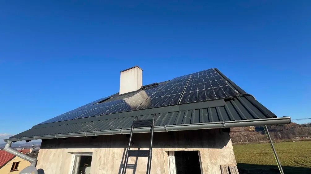 Instalacja paneli słonecznych na blaszanym dachu domu jednorodzinnego z drabina opartą o ścianę, widok z dołu na tle błękitnego nieba.