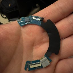 Marcin Fabicki - Uszkodzony, wygięty fragment obwodu drukowanego z czarnym elementem mocującym, trzymany w dłoni. Widoczne ślady uszkodzeń i brakujące elementy.