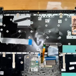 Wnętrze obudowy laptopa Lenovo po demontażu klawiatury, widoczne elementy elektroniki, taśmy połączeniowe i naklejki z datami produkcji podzespołów.